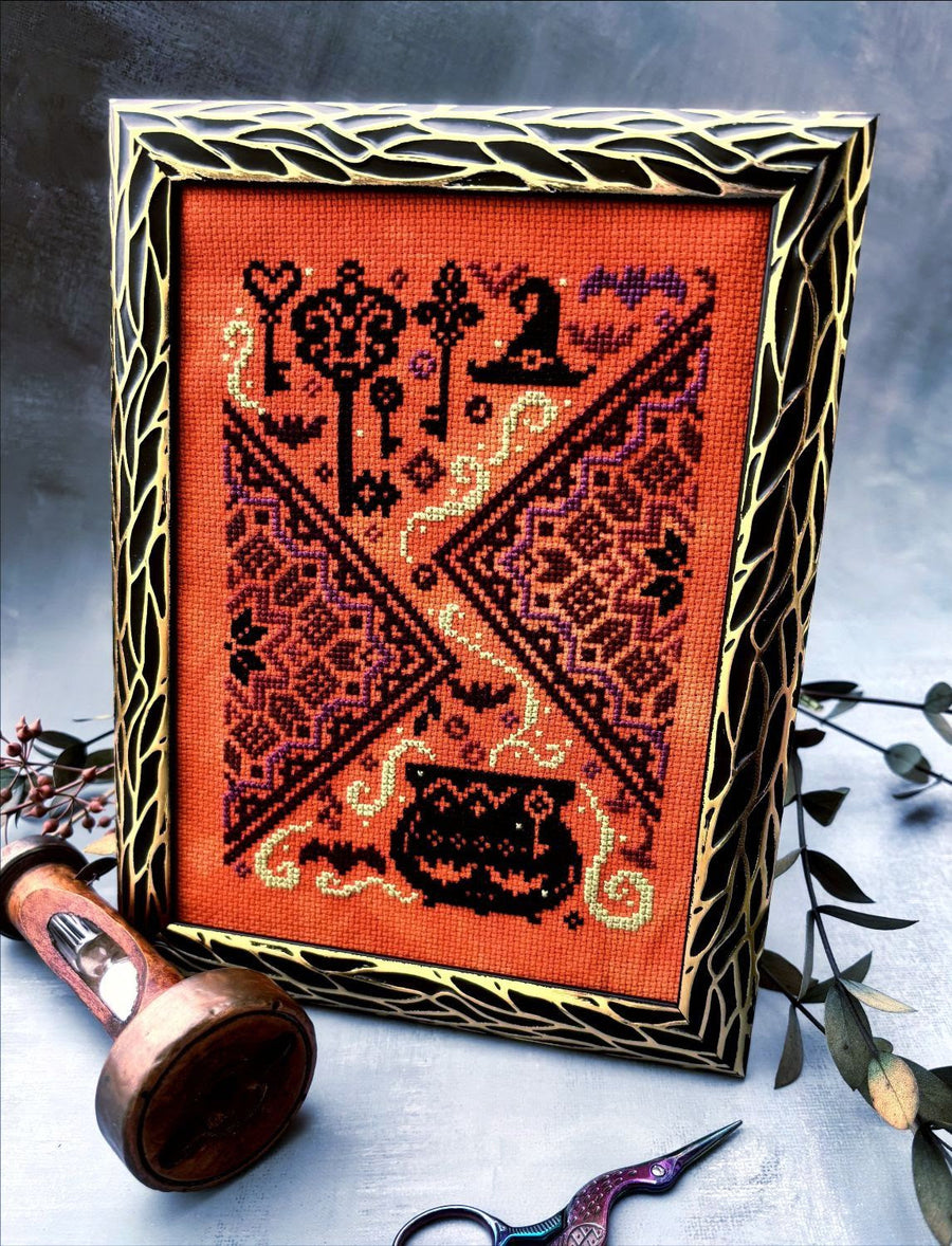 A framed cross-stitch artwork with a Halloween theme, featuring a bright orange fabric background, black and red embroidered motifs including keys, a witch hat, bats, and a cauldron, set in a decorative gold and black frame. The frame is styled on a gray surface with a wooden spool, embroidery scissors, and greenery.