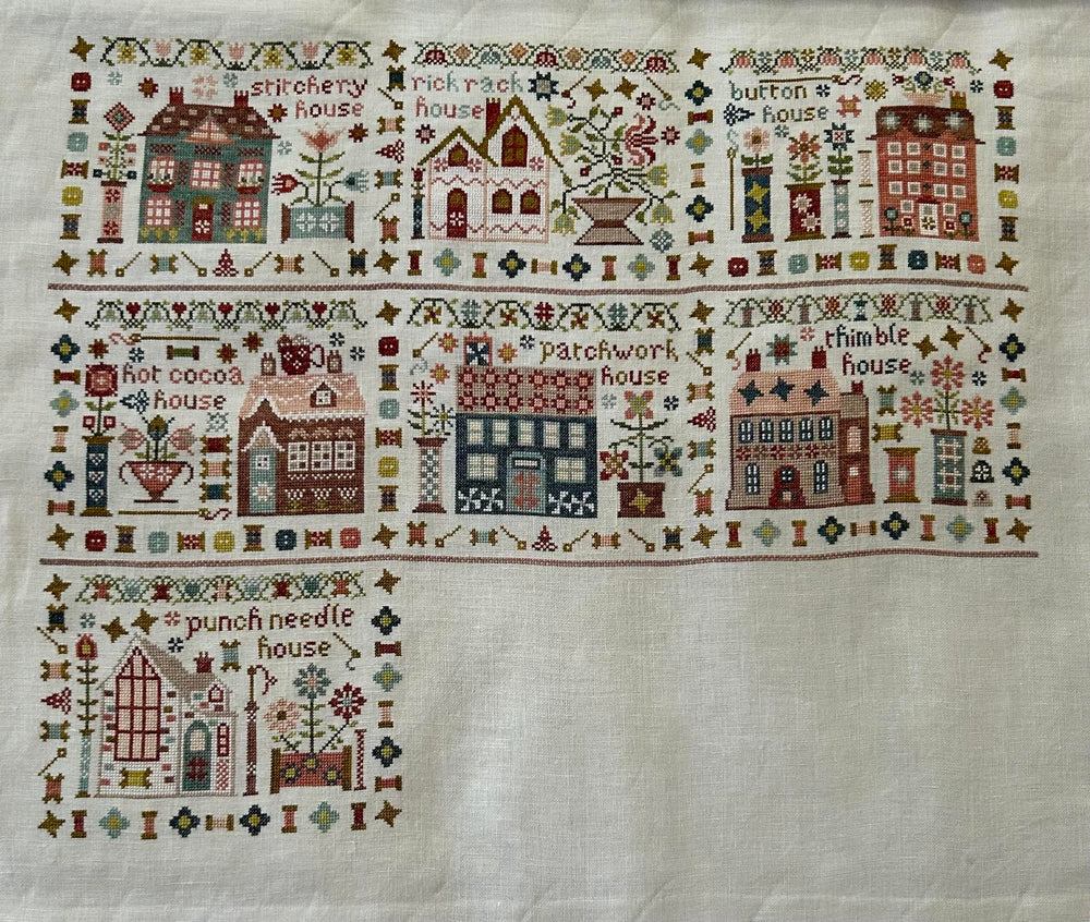 A square white needlepoint canvas featuring a stitched border of whimsical houses, trees, flowers, and decorative motifs, with a larger house design stitched in the lower left corner.