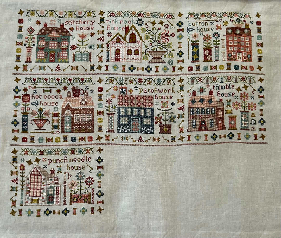 A square white needlepoint canvas featuring a stitched border of whimsical houses, trees, flowers, and decorative motifs, with a larger house design stitched in the lower left corner.