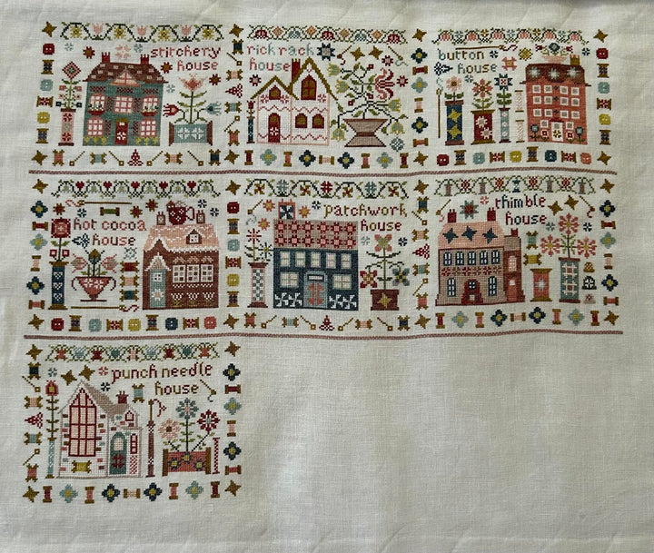 A square white needlepoint canvas featuring a stitched border of whimsical houses, trees, flowers, and decorative motifs, with a larger house design stitched in the lower left corner.