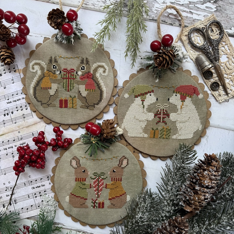 Three round embroidered cross-stitch ornaments featuring woodland animals holding red gifts, arranged on a white wooden surface with festive props including pinecones, red berries, song sheet, scissors, and evergreen branches.