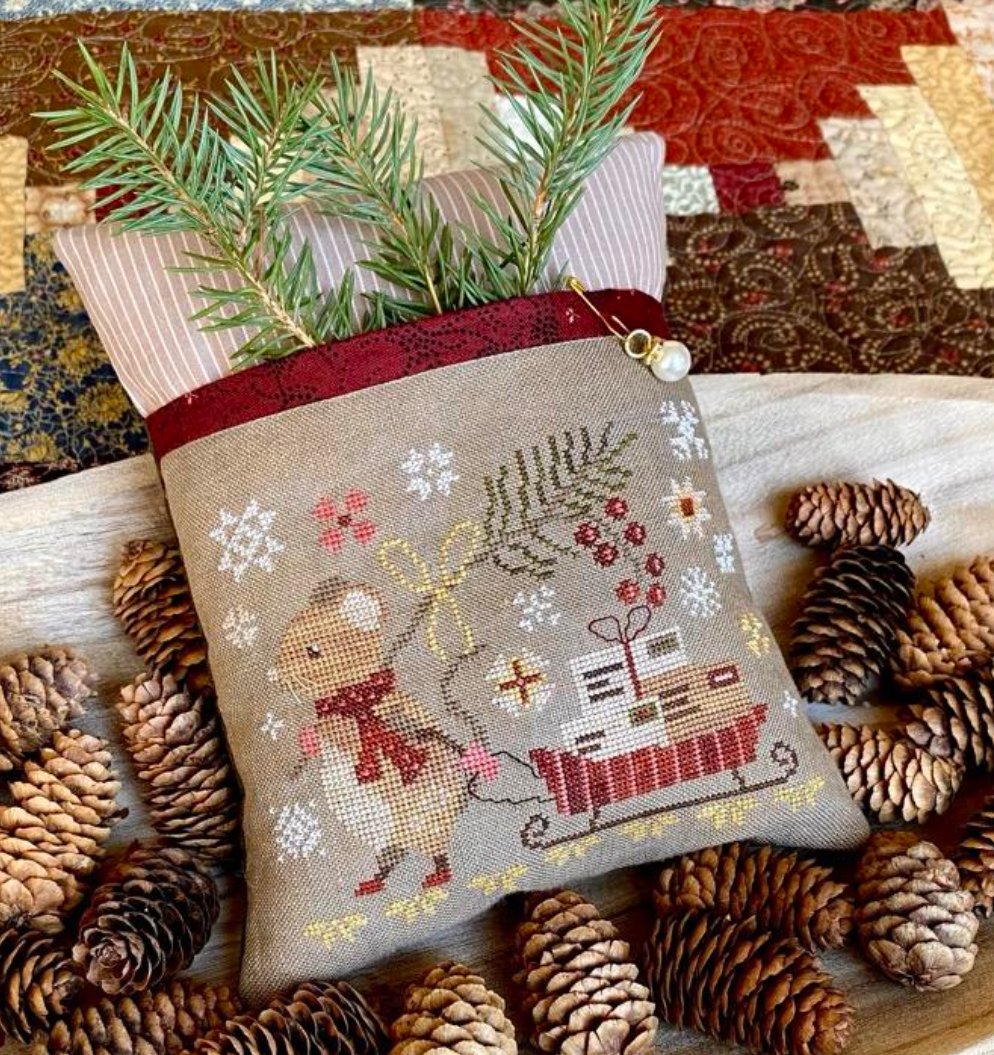 Decorative pillow with cross-stitch design of a reindeer and sleigh, surrounded by pine cones and a quilted fabric background.