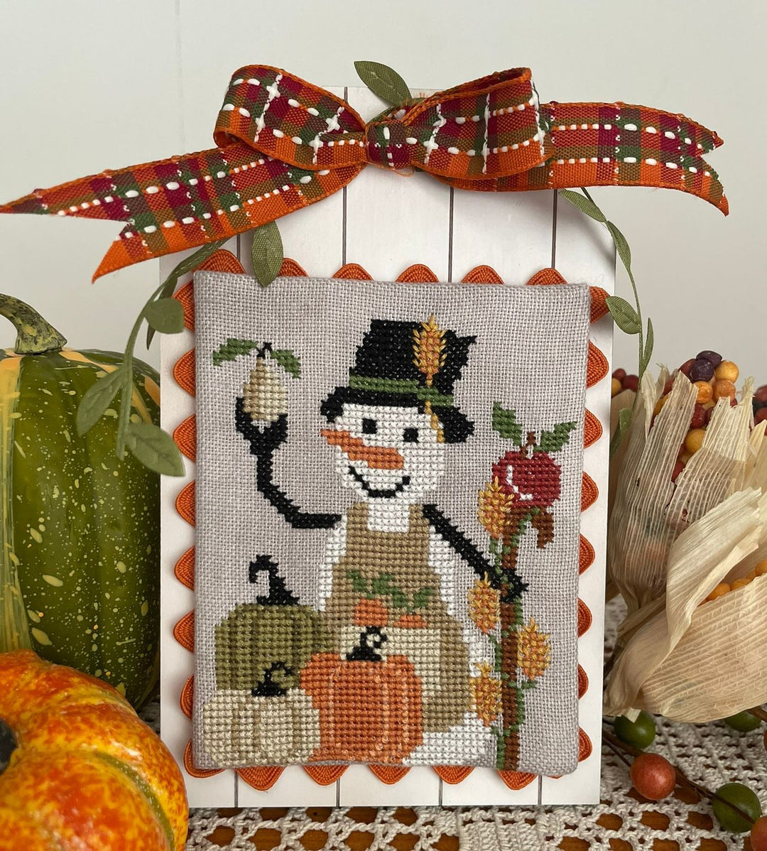 A framed cross-stitch artwork featuring a smiling snowman with a top hat and scarf, surrounded by pumpkins and an apple, displayed on a white picket-style board with an orange plaid bow. The scene is styled with autumn props including a green squash, dried corn husks, and small acorns.