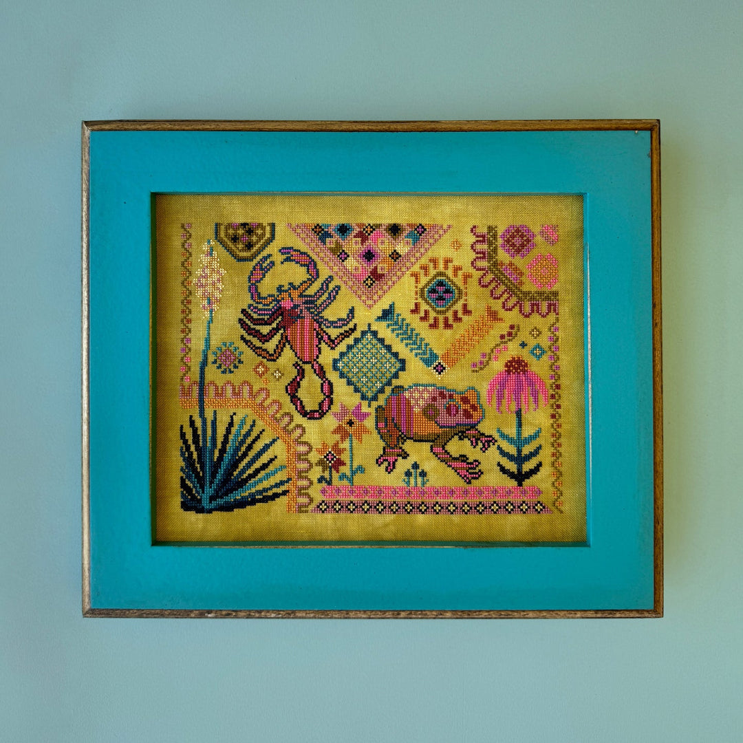 Framed cross-stitched artwork with a scorpion and frog design framed on a teal background