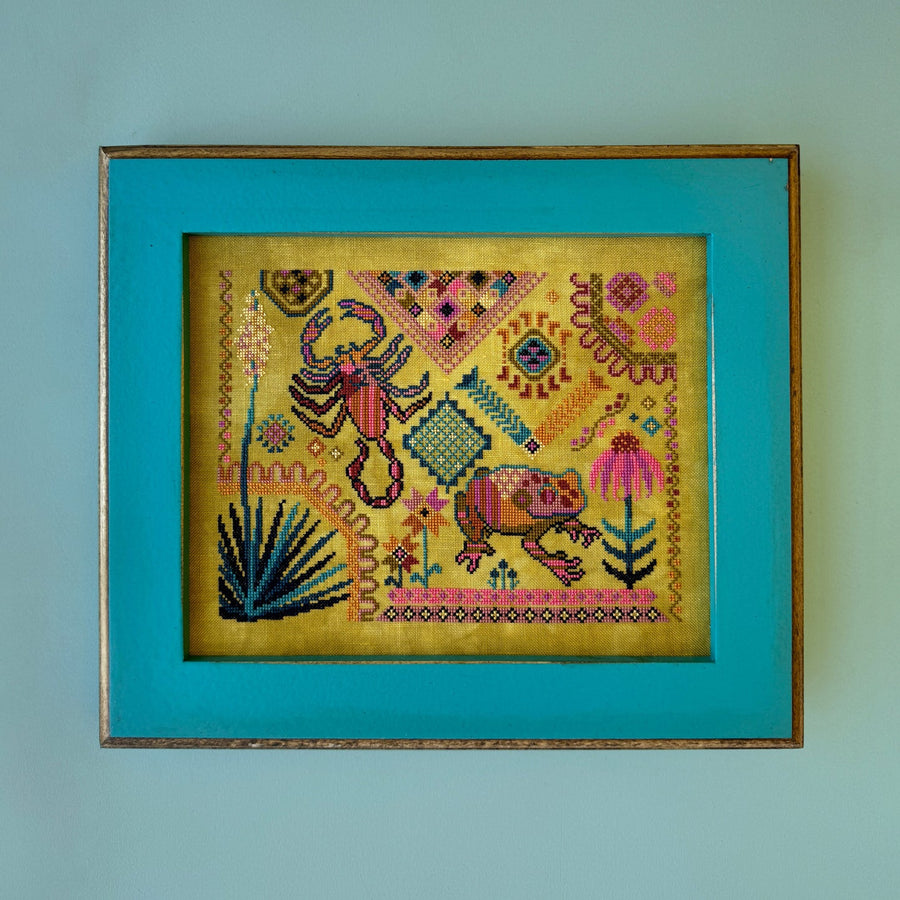 Framed cross-stitched artwork with a scorpion and frog design framed on a teal background
