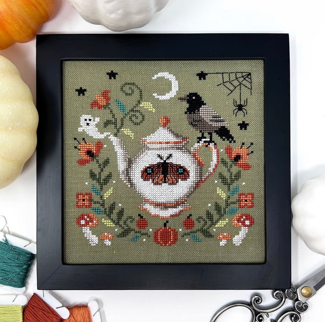 Framed cross-stitched artwork of a teapot with decorative elements on a white surface with pumpkins.