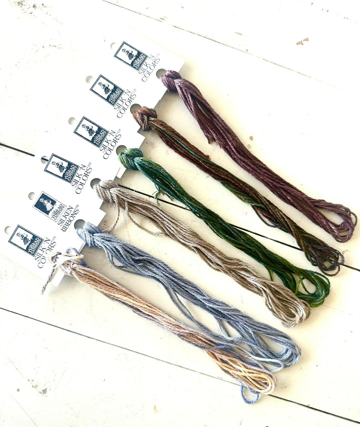 six skeins of embroidery floss in assorted earthy colors, including purple, green, beige, blue, and brown, attached to white paper labels from The Thread Gatherer displayed on a white wooden surface.