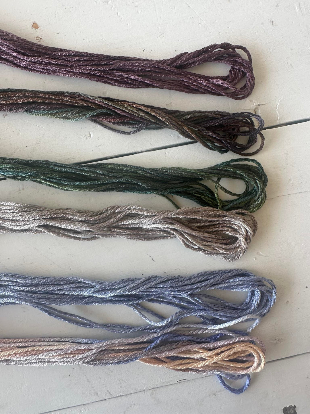 Six skeins of embroidery floss in muted earthy and cool tones, arranged vertically on a light off-white surface. The colors include plum, olive green, taupe, slate blue, and a light tan.