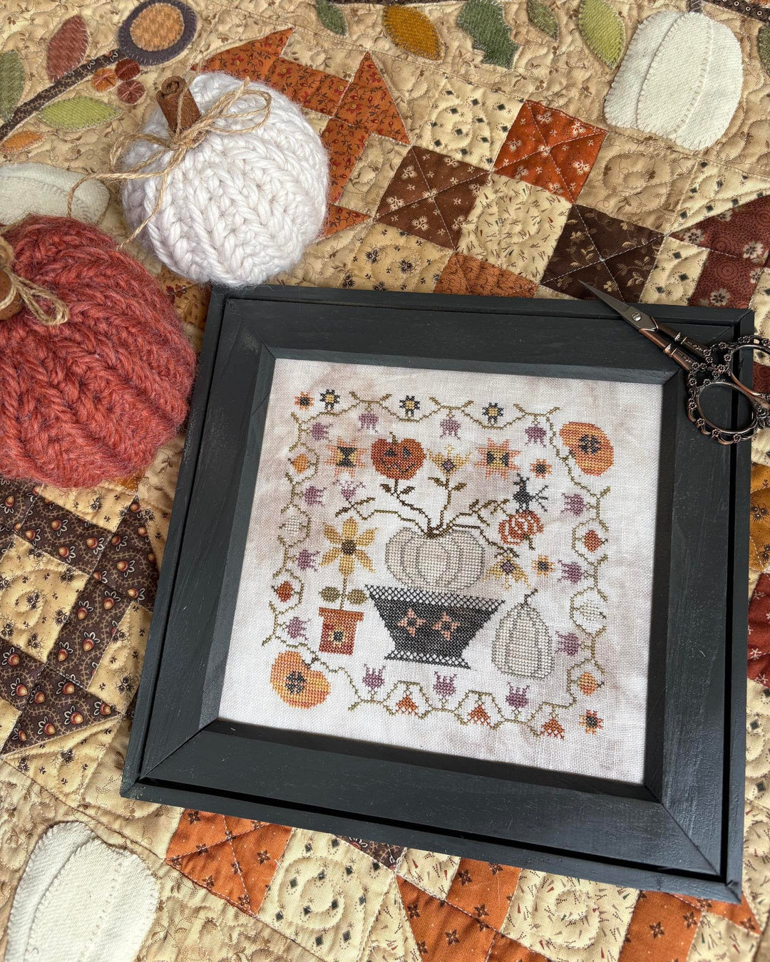 Framed cross-stitch pattern of pumpkins and flowers on a patchwork fabric background.