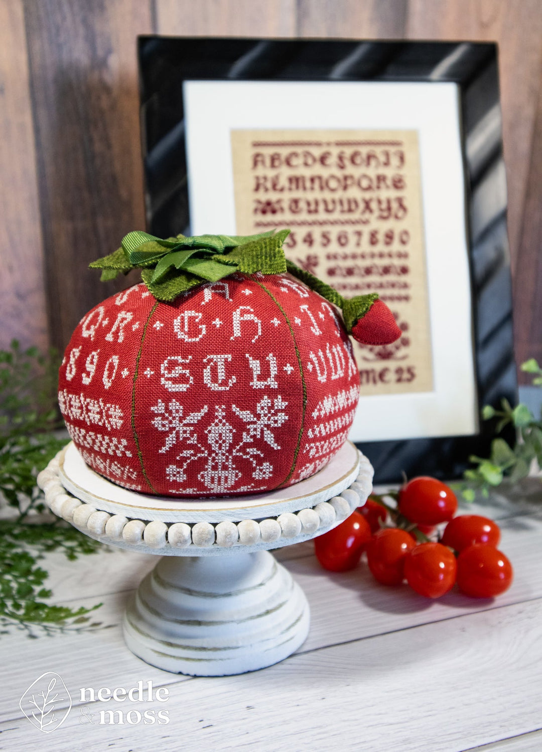 Decorative tomato-shaped pin cushion with cross-stitch pattern on a wooden surface, framed text in the background.