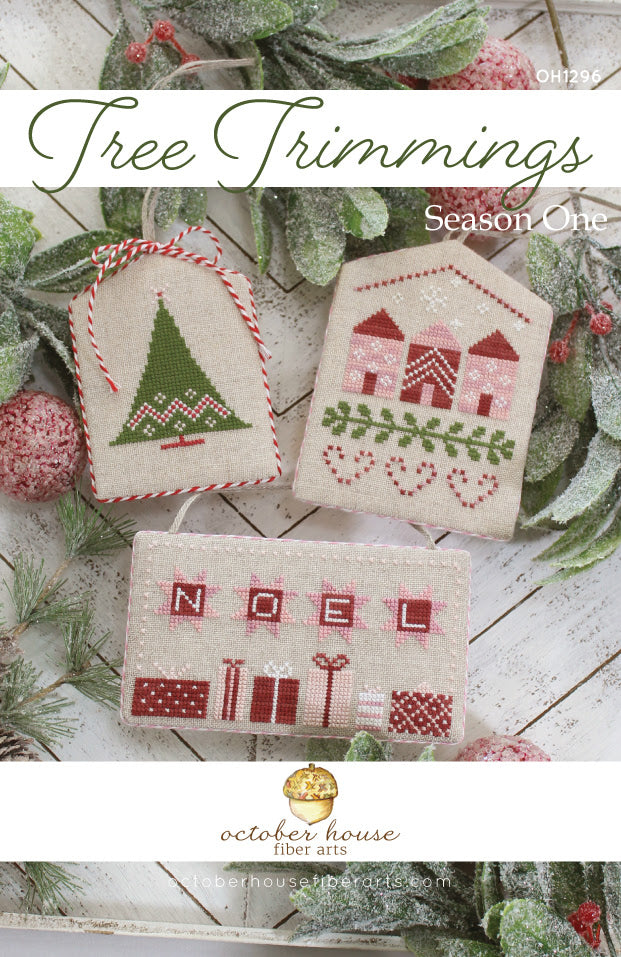 Cross-stitched Christmas ornaments with 'NOEL' and house designs on a wooden surface with greenery.