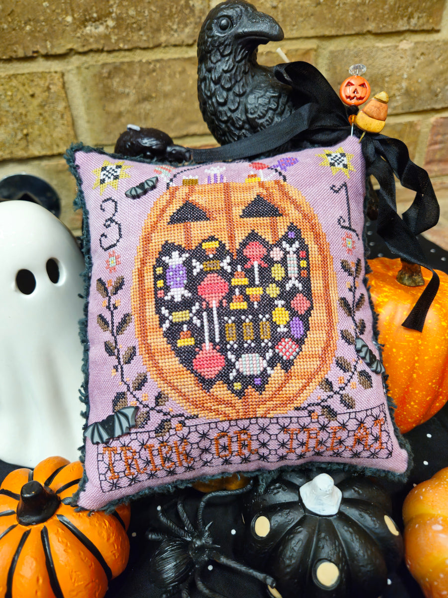A square lavender cross-stitch pillow featuring a stitched jack-o'-lantern with colorful mushrooms and bats, surrounded by Halloween-themed props including small pumpkins, a black raven figurine, a white ghost, and a brick wall background.