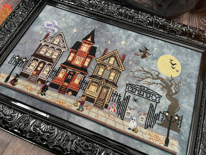 A framed needlepoint or cross-stitch artwork depicting three haunted Victorian houses at night, with a full moon, bats, a bare tree, and a gate labeled 'WELCOME'. The piece is enclosed in an ornate black frame and photographed on a brown fabric surface.