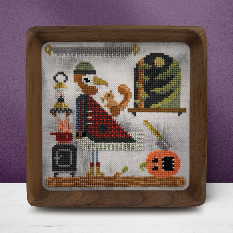 A framed cross-stitch artwork featuring a stylized turkey in a red plaid shirt next to a green-and-black striped bird, with autumn-themed elements including a pumpkin, lantern, and axe on a white fabric background, all enclosed in a square wooden frame against a purple wall.