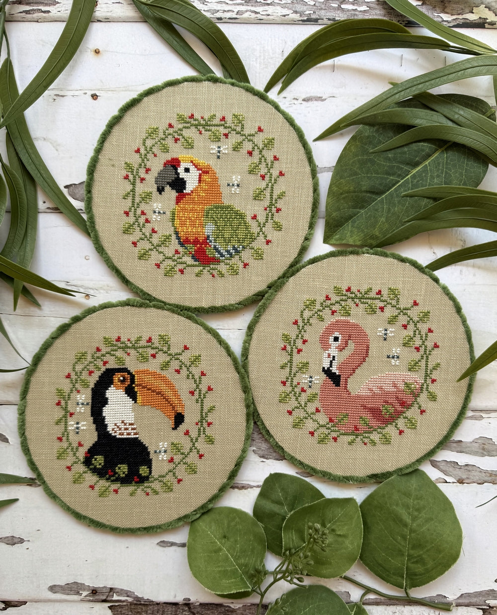 Three cross-stitched roundels featuring a parrot, toucan, and flamingo on a wooden surface with leaves.