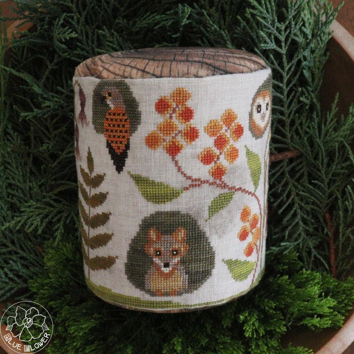 A cylindrical fabric-covered needlepoint canvas featuring woodland animal and floral motifs, displayed on a bed of green pine branches. The design includes a hedgehog, fox, owls, berries, and leaves on an off-white background.