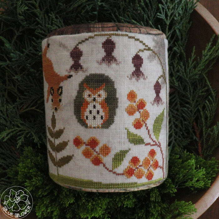 A cylindrical woven textile item, likely a small basket, bag, or decorative container, featuring a cross-stitch style woodland design with an owl, fox, hanging flowers, and berries. The item is displayed upright on green evergreen foliage.