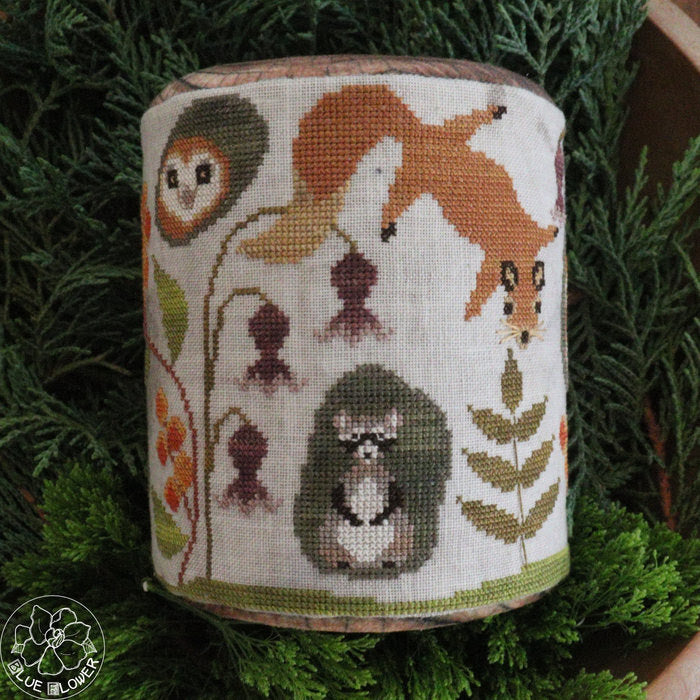 A cylindrical needlepoint canvas featuring woodland animal and floral designs, displayed on a bed of green pine branches. The design includes a fox, hedgehog, owl, and flowers on an off-white background.