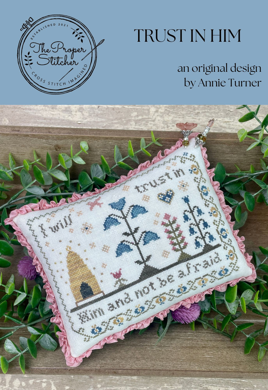 Cross-stitched pillow with floral design and text, surrounded by greenery on a wooden surface.