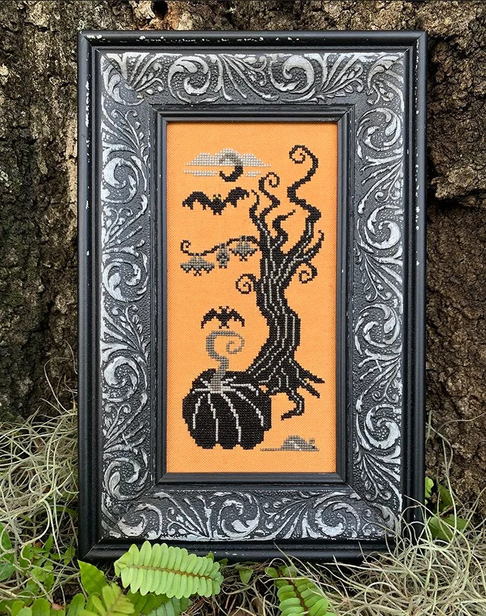 Framed cross-stitched artwork of a Halloween-themed design with bats and a pumpkin on an orange background.