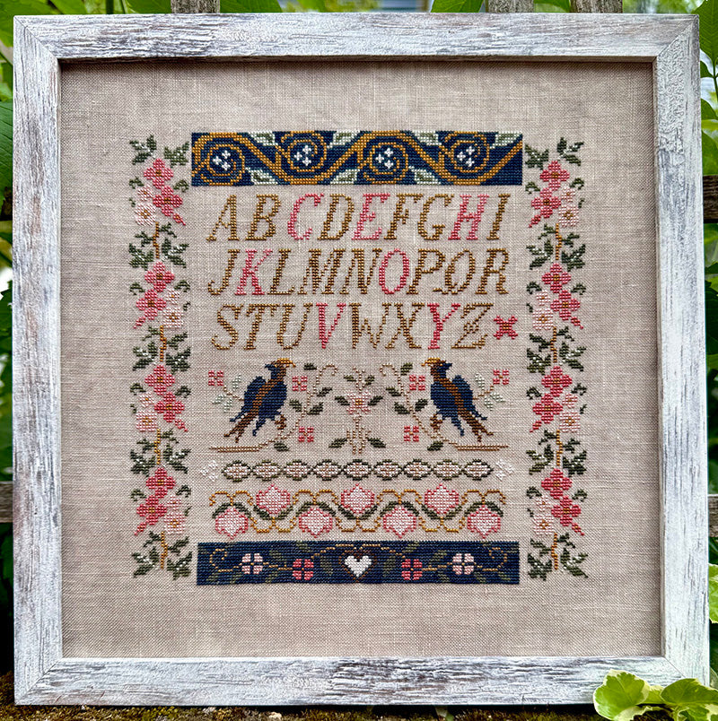 A framed cross-stitch sampler featuring the alphabet, two birds, floral borders, and decorative geometric patterns on a beige linen background, enclosed in a distressed white wooden frame.