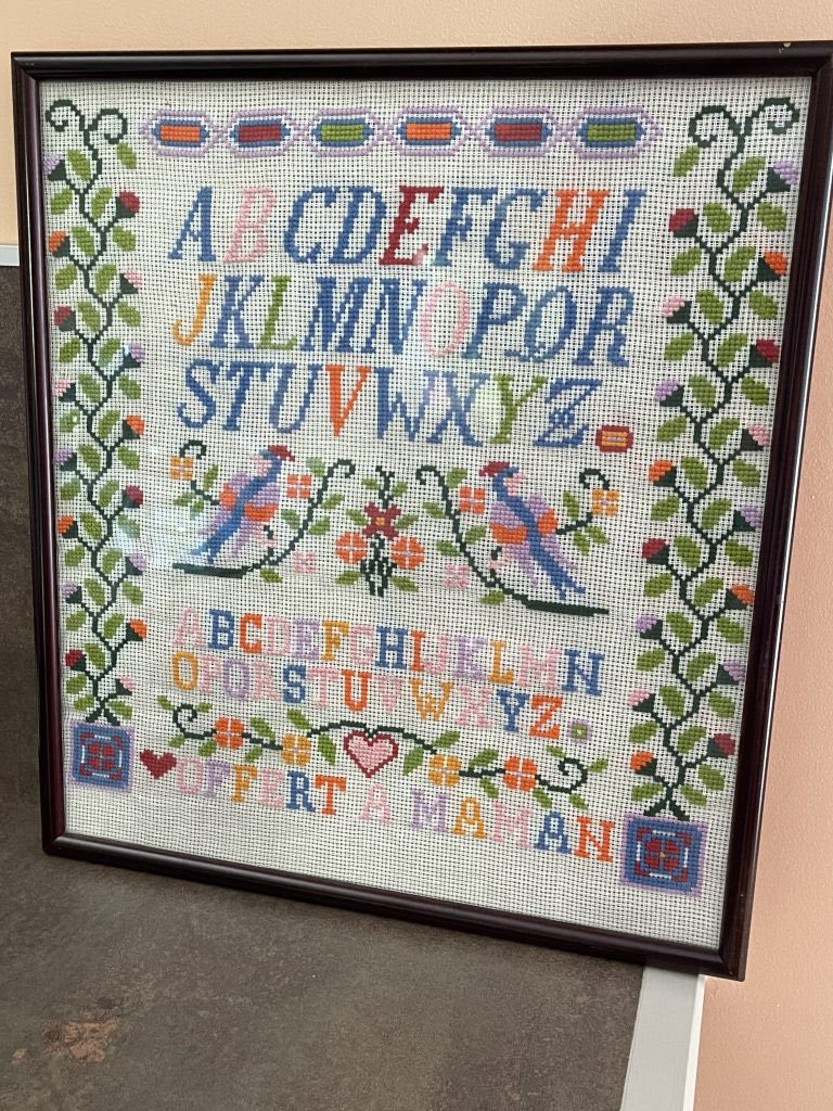 A framed cross-stitch sampler featuring the alphabet in two rows, two blue birds, floral motifs, and decorative borders, photographed leaning against a wall on a tabletop.