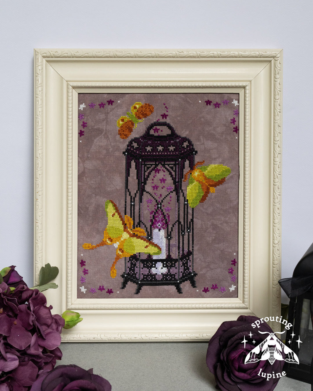 Framed cross-stitch artwork featuring a black birdcage with a white candle and yellow butterflies on a mauve background, displayed in an ornate cream frame with decorative flowers in the foreground.