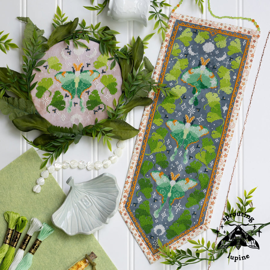 A styled flat-lay of needlework/cross-stitch patterns or finished textile pieces featuring green moths on a gray background, surrounded by faux greenery, thread spools, a shell dish, and a white paneled backdrop. A visible watermark reads 'needlework lupine'.