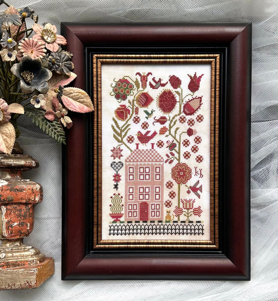 Framed cross-stitched artwork with a house and floral design on a textured surface.