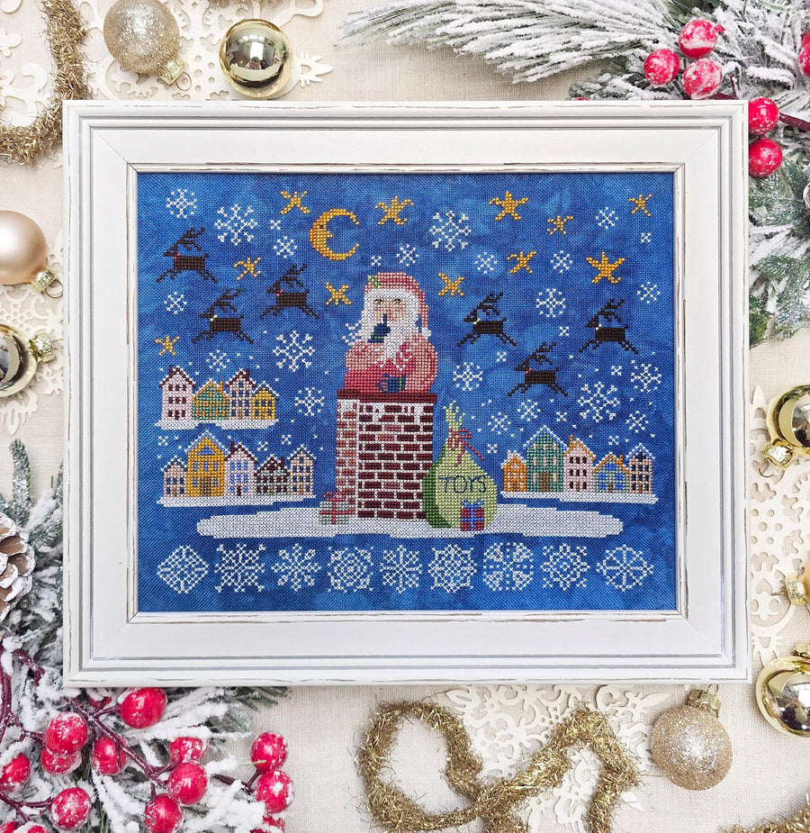 A framed Christmas needlework or cross-stitch artwork featuring Santa Claus standing on a brick chimney with a snowy village, reindeer, snowflakes, and stars on a blue background, displayed in a white frame with holiday decorations around it.