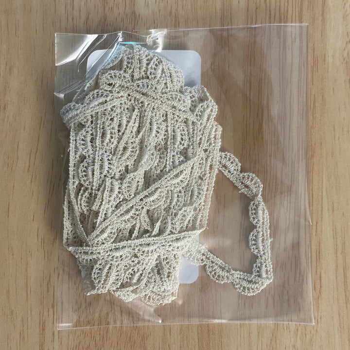 A piece of off-white floral lace trim packaged in a clear plastic bag, placed on a light wood grain surface.