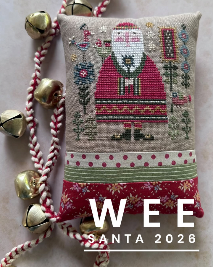 A square embroidered pillow featuring a festive Santa Claus design on a beige linen background, with red-and-white braided cord and gold bells draped around it. Large white text at the bottom reads 'WEE SANTA 2026'.