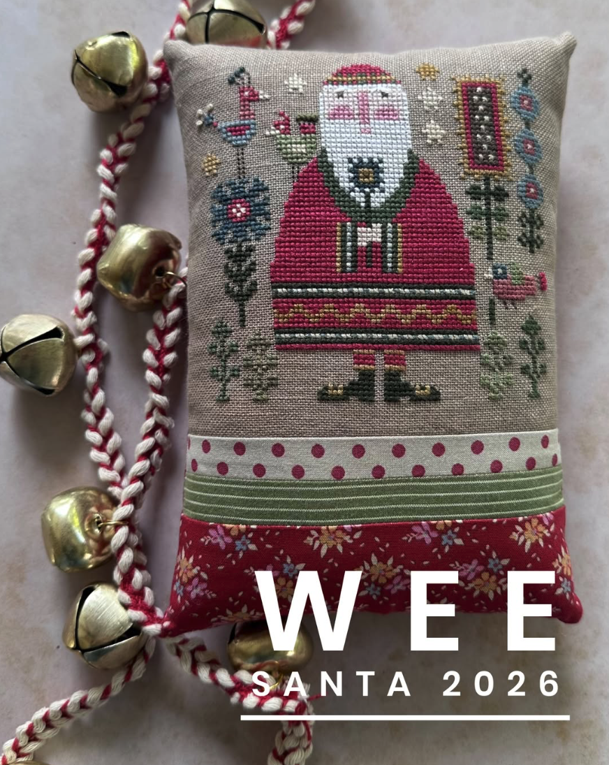 A square embroidered pillow featuring a festive Santa Claus design on a beige linen background, with red-and-white braided cord and gold bells draped around it. Large white text at the bottom reads 'WEE SANTA 2026'.