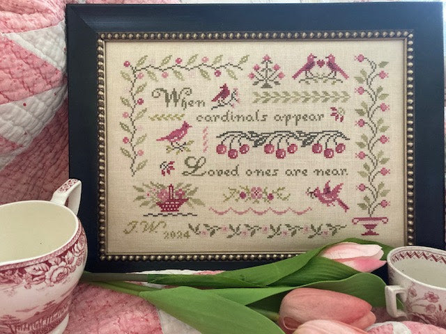 Framed cross-stitched piece with floral and bird patterns, placed on a surface with tulips and a teacup.