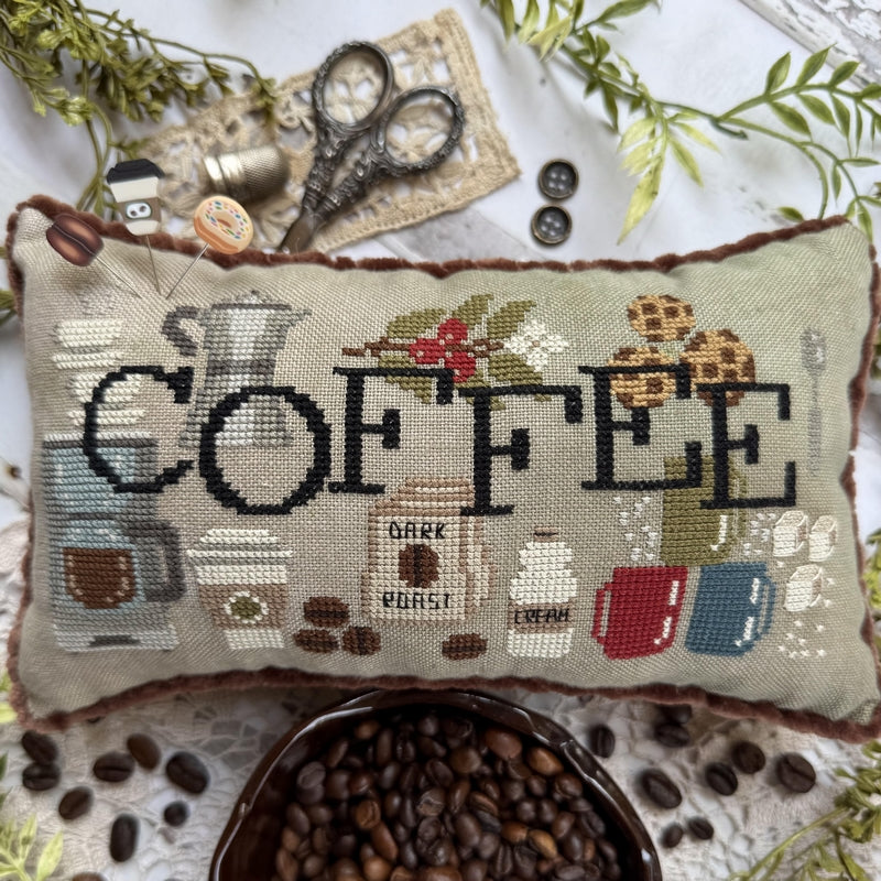 A rectangular cross-stitch pillow featuring the word 'COFFEE' in black thread with illustrated coffee-themed motifs including mugs, a coffee pot, beans, and pastries. The pillow is styled on a white lace surface with scattered coffee beans, scissors, buttons, and decorative greenery.
