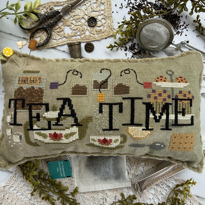 A rectangular needlepoint cushion with a 'TEA TIME' design, styled in a flat-lay arrangement with tea accessories, scissors, and lace on a white background.