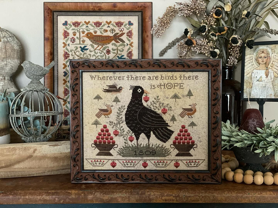 Framed cross-stitched artwork with a crow and text on a shelf with decorative items.