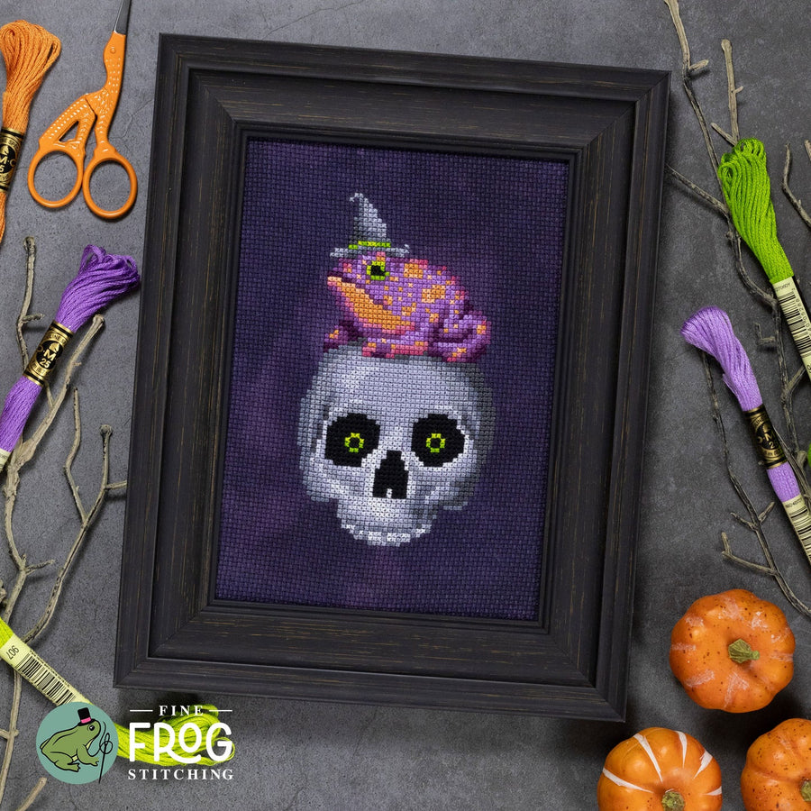 A framed cross-stitch artwork featuring a white skull with green eyes and a pink frog wearing a purple witch hat on a dark purple background, styled on a gray surface with embroidery floss, scissors, pumpkins, and branches. The Fingertip Frog Stitching logo appears in the lower left corner.