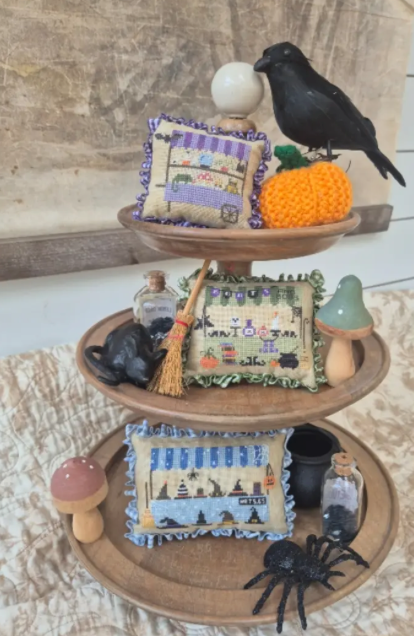 A three-tier wooden display stand styled with Halloween-themed decorative items, including embroidered pillows, a black raven figurine, a crocheted orange pumpkin, miniature bottles, a broom, mushrooms, a black spider, and a white orb.