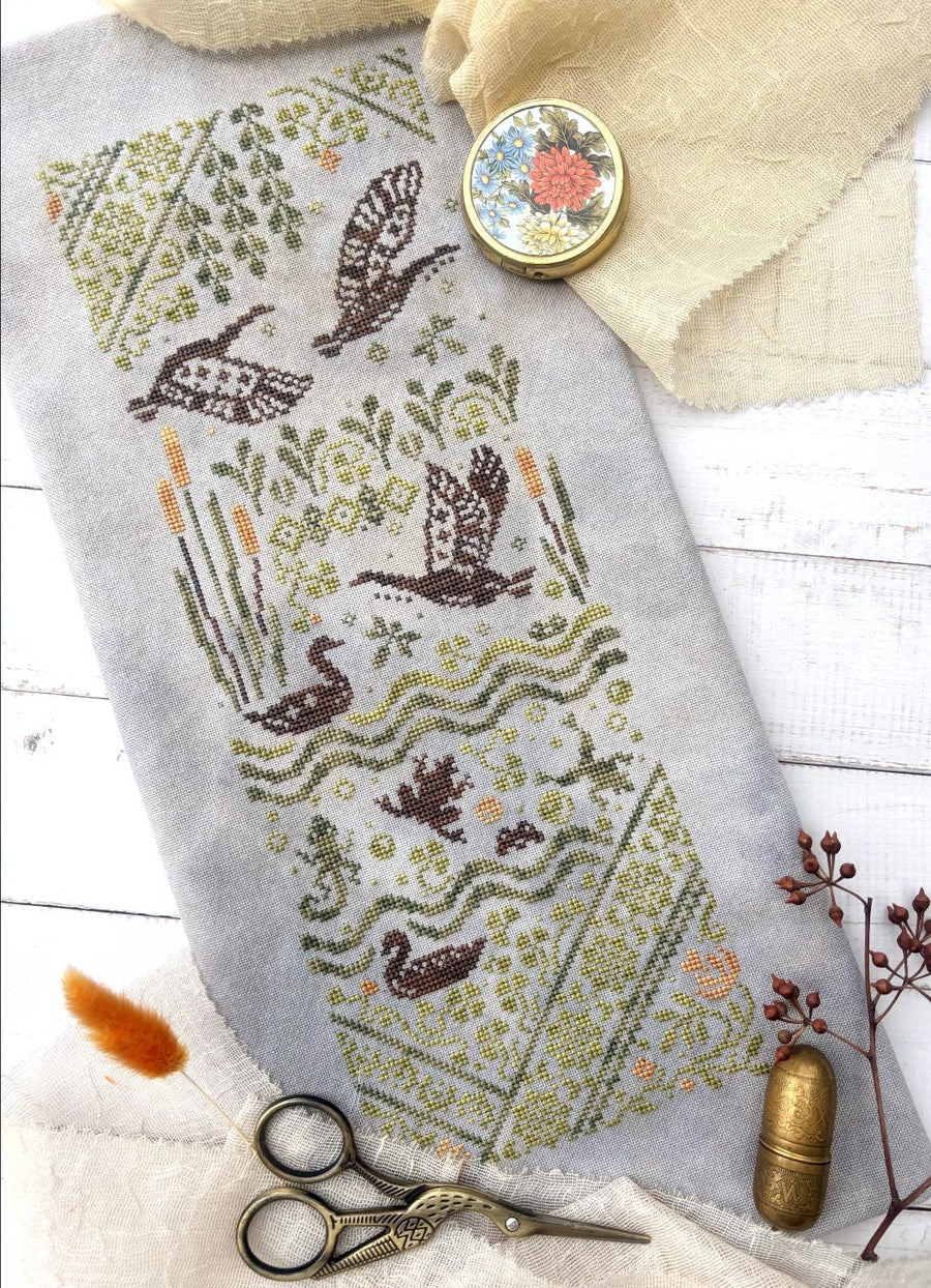 A needlepoint canvas featuring a nature pond scene with ducks, reeds, and birds, styled in a flat-lay with scissors, fabric, and decorative items on a white wood background.