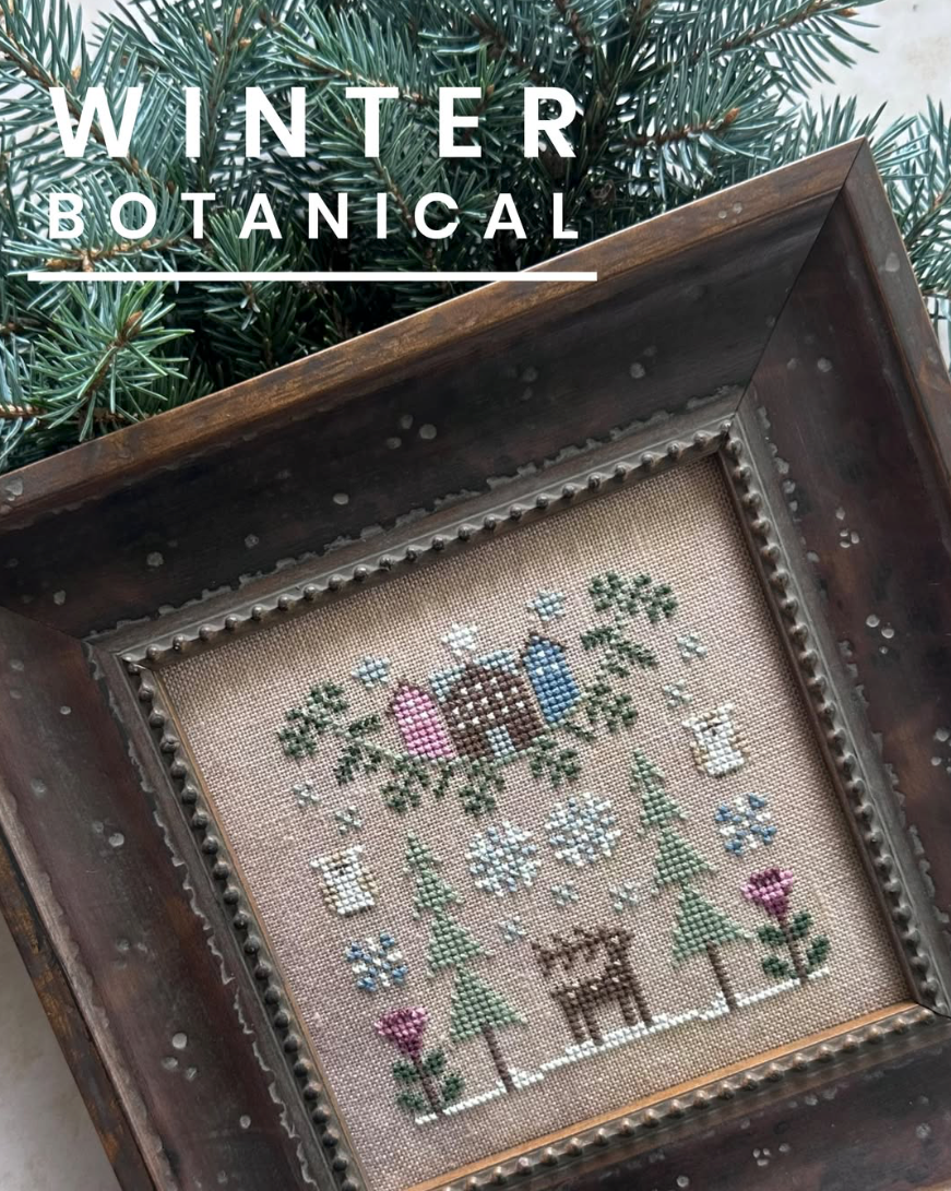 A framed cross-stitch artwork titled 'Winter Botanical' featuring a snowy winter scene with a deer, evergreen trees, flowers, and birds on a beige fabric background, set inside a dark wooden frame with pine branches behind it.