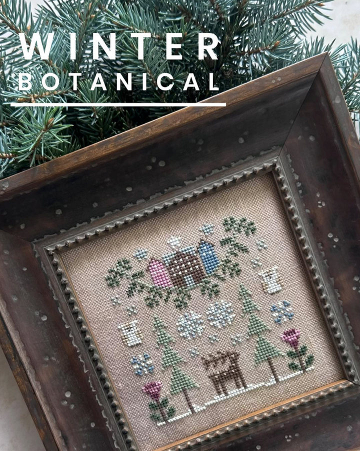 A framed cross-stitch artwork titled 'Winter Botanical' featuring a snowy winter scene with a deer, evergreen trees, flowers, and birds on a beige fabric background, set inside a dark wooden frame with pine branches behind it.