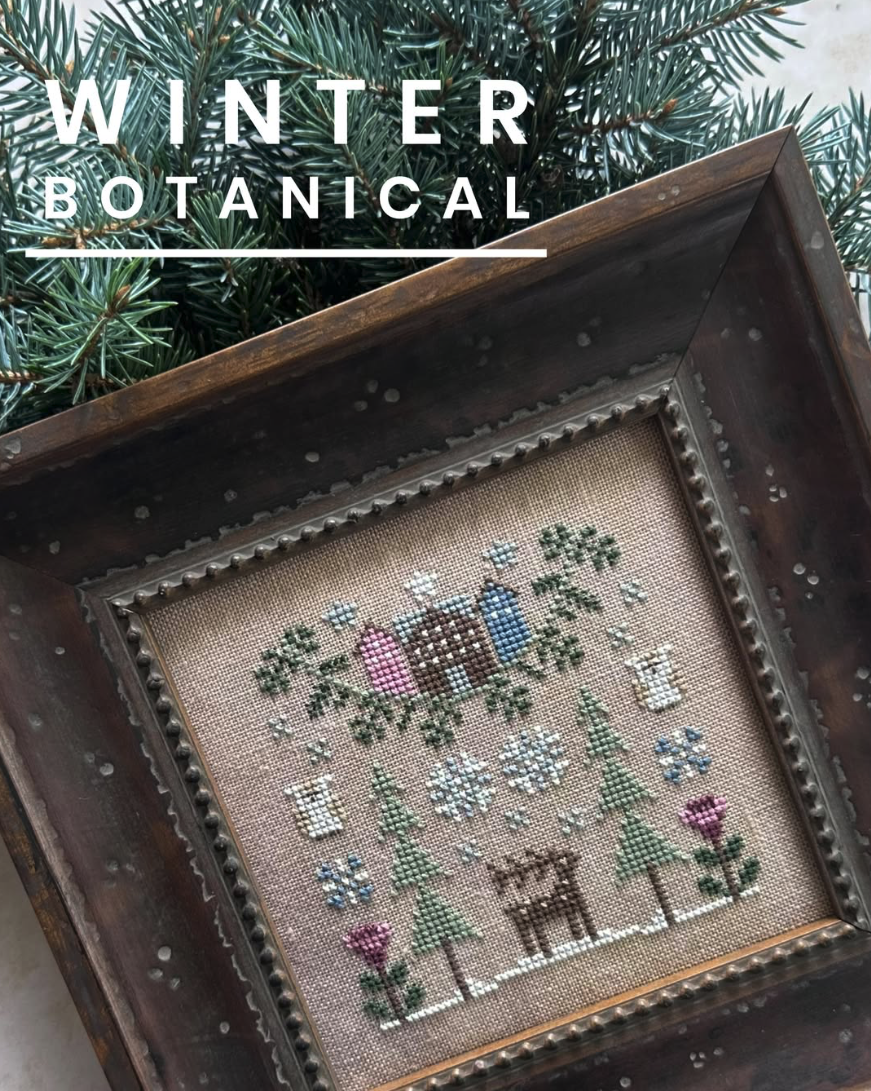 A framed cross-stitch artwork titled 'Winter Botanical' featuring a snowy winter scene with a deer, evergreen trees, flowers, and birds on a beige fabric background, set inside a dark wooden frame with pine branches behind it.