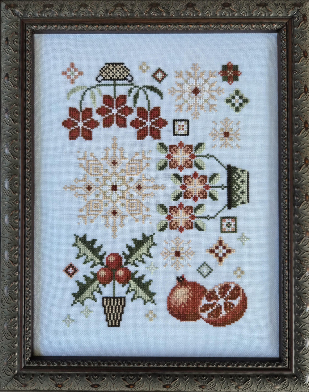 A framed cross-stitch artwork featuring winter motifs including snowflakes, flowers, holly, berries, and pomegranates on a white fabric background, surrounded by an ornate dark brown frame.