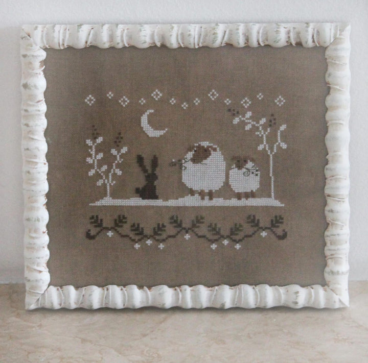A framed cross-stitch artwork featuring a bunny, sheep, bird, crescent moon, and delicate floral motifs on a taupe fabric background, surrounded by a white ruffled frame.