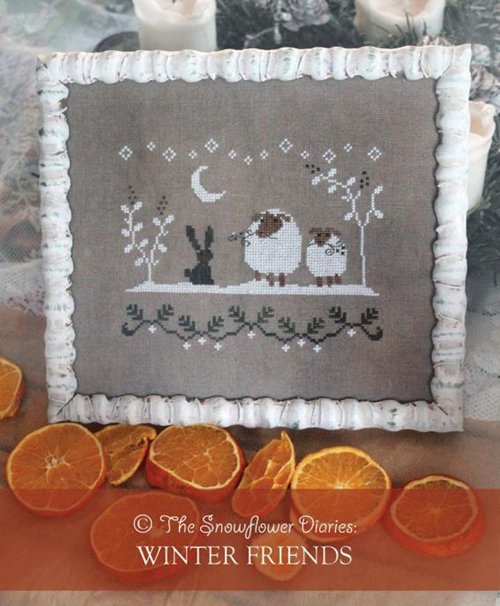 A square cross-stitch franed design with a white frame, featuring a winter scene with a crescent moon, stars, a rabbit, and two sheep on a taupe fabric background. The pillow is styled with dried orange slices and candles.