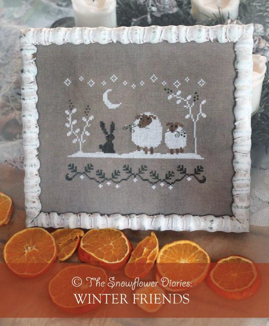 A square cross-stitch franed design with a white frame, featuring a winter scene with a crescent moon, stars, a rabbit, and two sheep on a taupe fabric background. The pillow is styled with dried orange slices and candles.