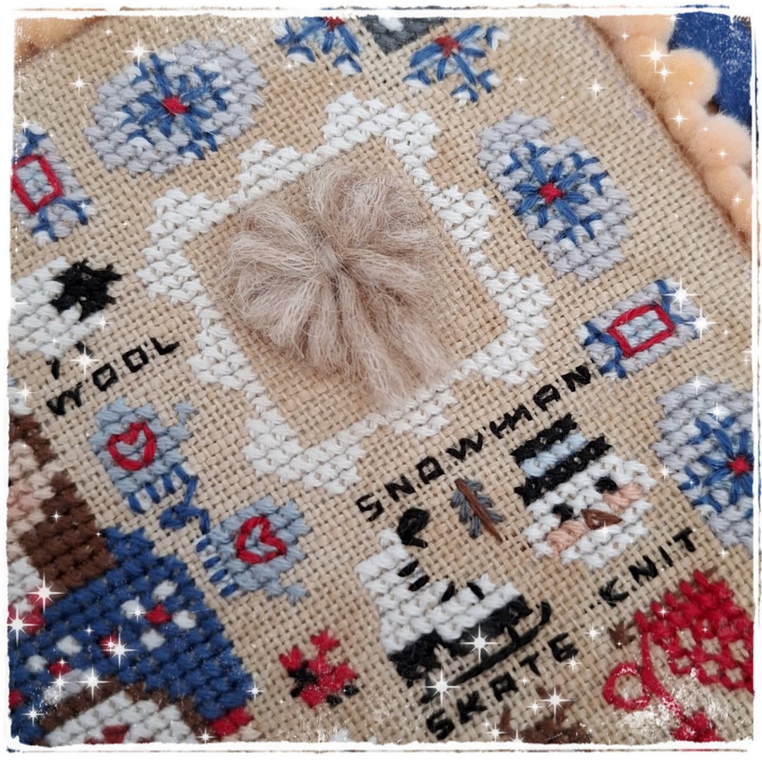 A close-up of a handmade cross-stitch or needlepoint textile featuring a snowman design, with a fluffy beige pom-pom attached in the center. The fabric has a warm beige base with stitched details in white, blue, red, black, and brown, surrounded by a decorative sparkly border.
