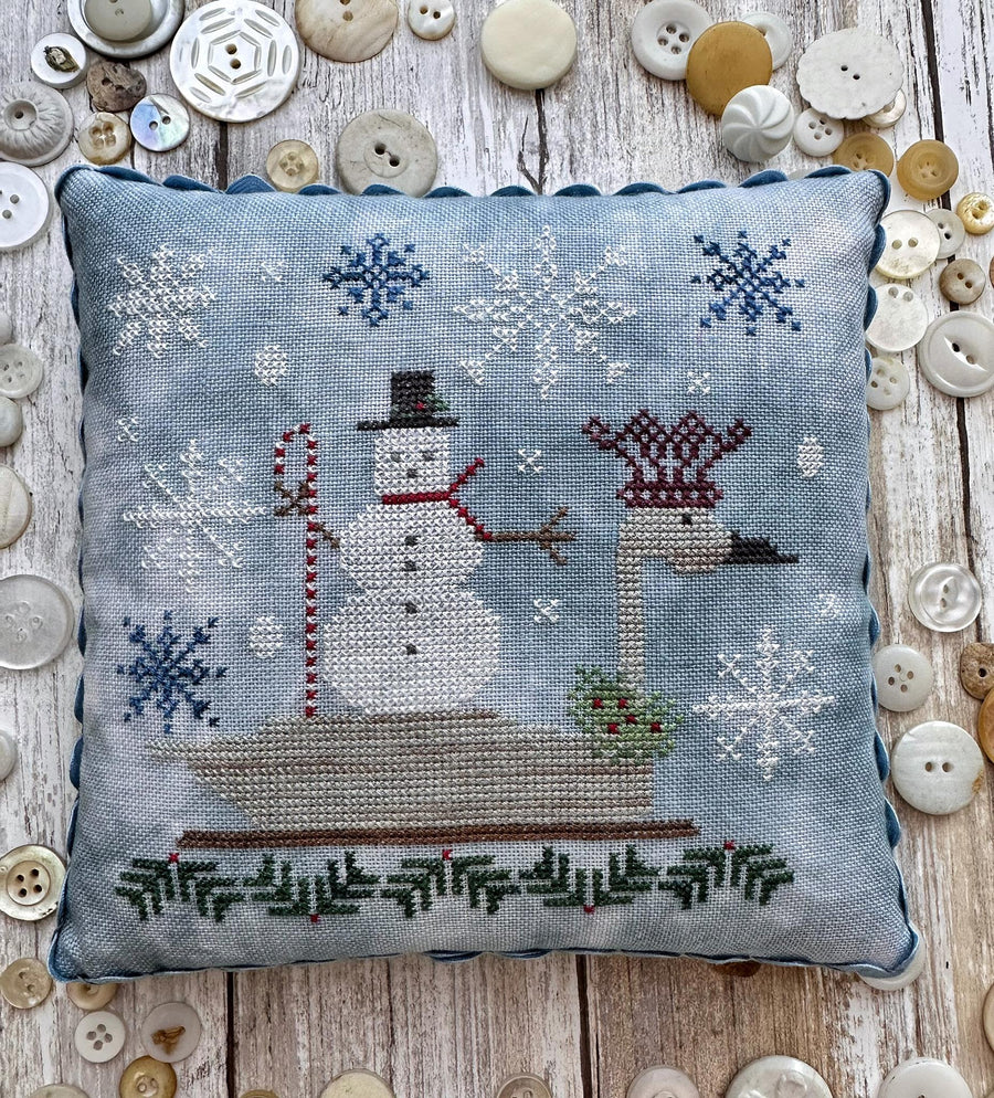 A square cross-stitch pillow with a winter scene featuring a snowman on a sled and a goose, surrounded by scattered decorative buttons on a rustic wood background.