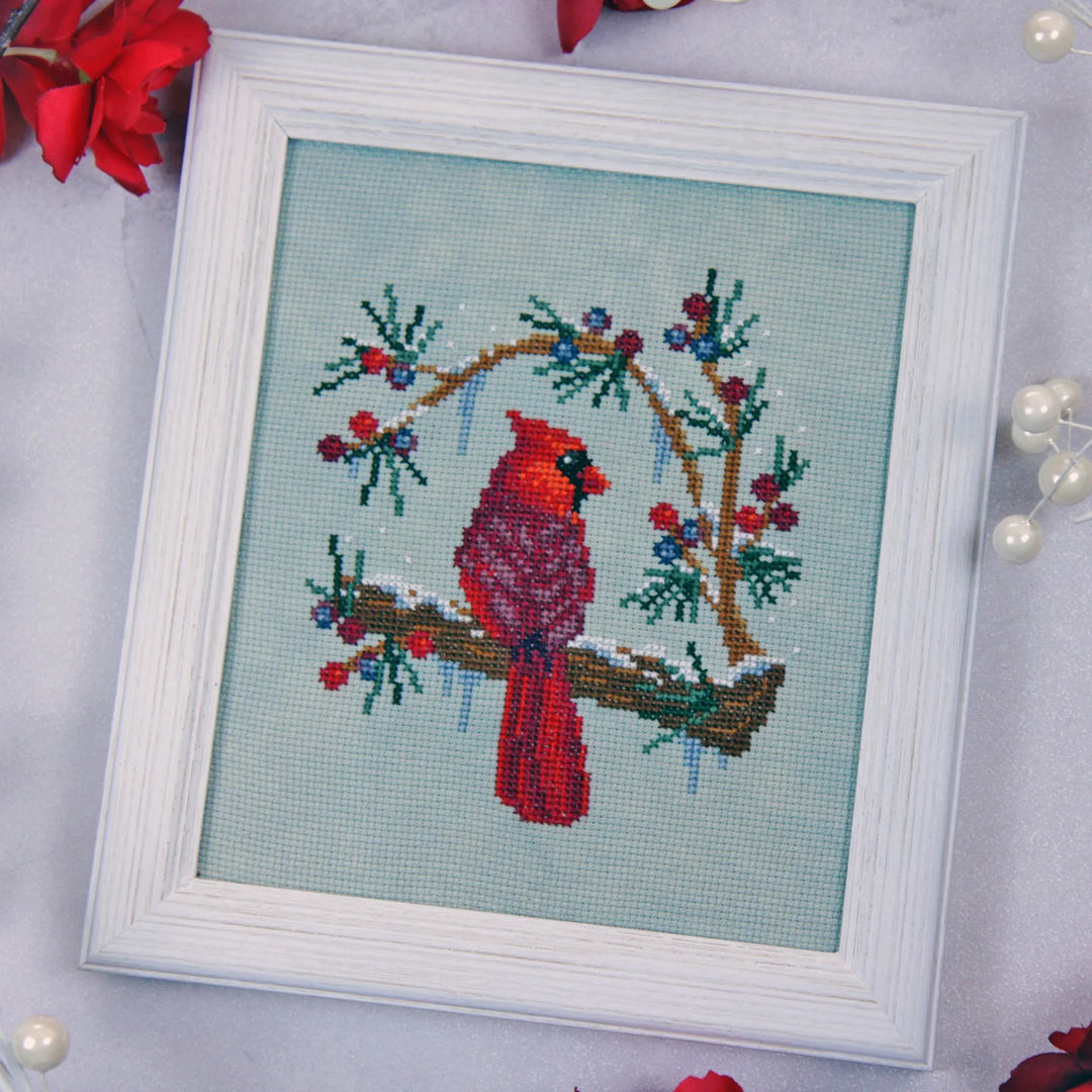 A framed cross-stitch artwork featuring a red cardinal perched on a snowy branch with blue berries, set in a white frame on a light background with decorative red flowers and pearl beads.